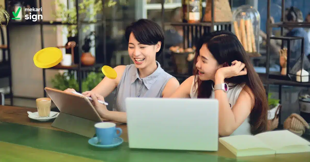 Dua pekerja freelance berdiskusi sambil menggunakan laptop dan tablet di kafe, mendukung kolaborasi kerja digital dengan Mekari Sign